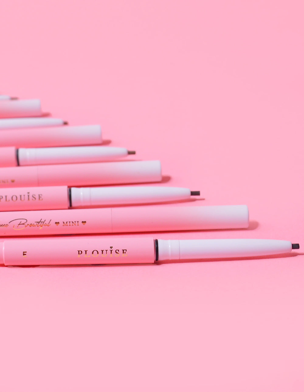 A collection of P.Louise Mini Eyebrow Pencils laid in a row with the brand name visible on the body of the pencils, displayed against a pink background