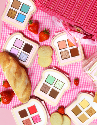 A set of mini sandwich-style eyeshadow palettes with various shades of makeup colours displayed on a pink gingham surface, accompanied by strawberries and a piece of bread.
