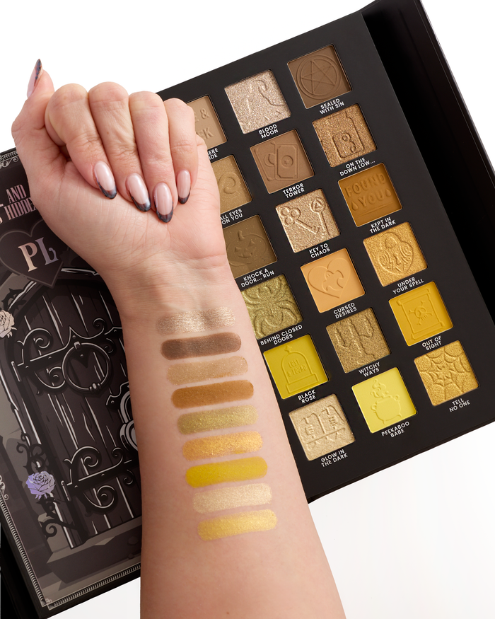 A close-up of a hand and forearm swatched with makeup from an open halloween eyeshadow palette. The palette contains 18 square pans in warm metallic and matte shades of gold, bronze, brown, and yellow, with decorative gothic lettering and illustrations.