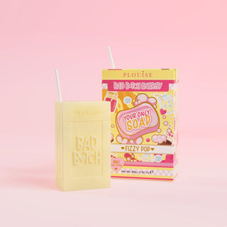 A yellow bar of soap with 'Bad Bitch' engraved on it, accompanied by a colourful juice box package with 'Fizzy Pop' labelled on it, presented on a pink background.
