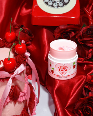 A container of P.Louise Plot Twist Make It Melt Makeup Cleansing Balm displayed on a red fabric surface with decorative elements including a cherry and a vintage red phone in the background.