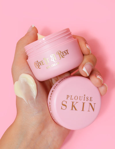 A person holding a pink jar of P.Louise Give It A Rest eye cream with the lid off, showing the product name and branding on the jar.
